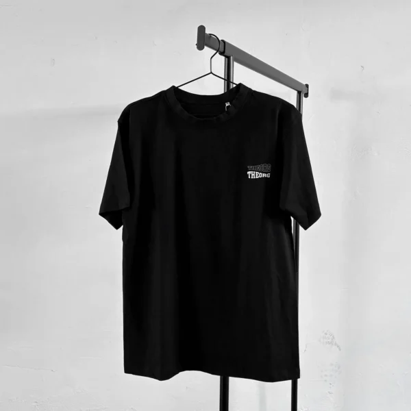 Schwarzes T-Shirt auf an einem Kleiderbügel an einer Kleiderstange: Frontdruck: zwei wellenförmige THEORG-Schriftzüge oben mit einer weißen Konturlinie unten weiß ausgefüllt.