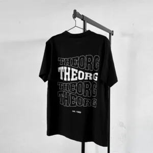 Schwarzes T-Shirt auf an einem Kleiderbügel an einer Kleiderstange. Der Rückendruck ist viermal THEORG in einer wellenförmigen Schrift untereinander. Das zweite THEORG von oben ist weiß ausgefüllt, die anderen haben eine weißen Konturlinie. Darunter steht est. 1989.