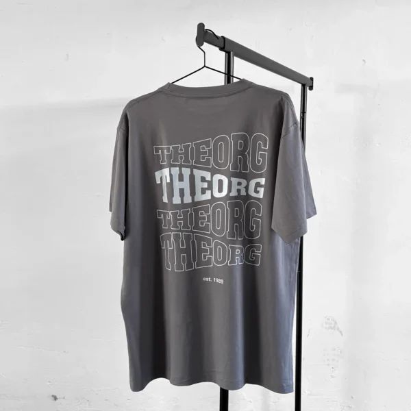 Graues T-Shirt auf an einem Kleiderbügel an einer Kleiderstange. Der Rückendruck ist viermal THEORG in einer wellenförmigen Schrift untereinander. Das zweite THEORG von oben ist weiß ausgefüllt, die anderen haben eine weißen Konturlinie. Darunter steht est. 1989.